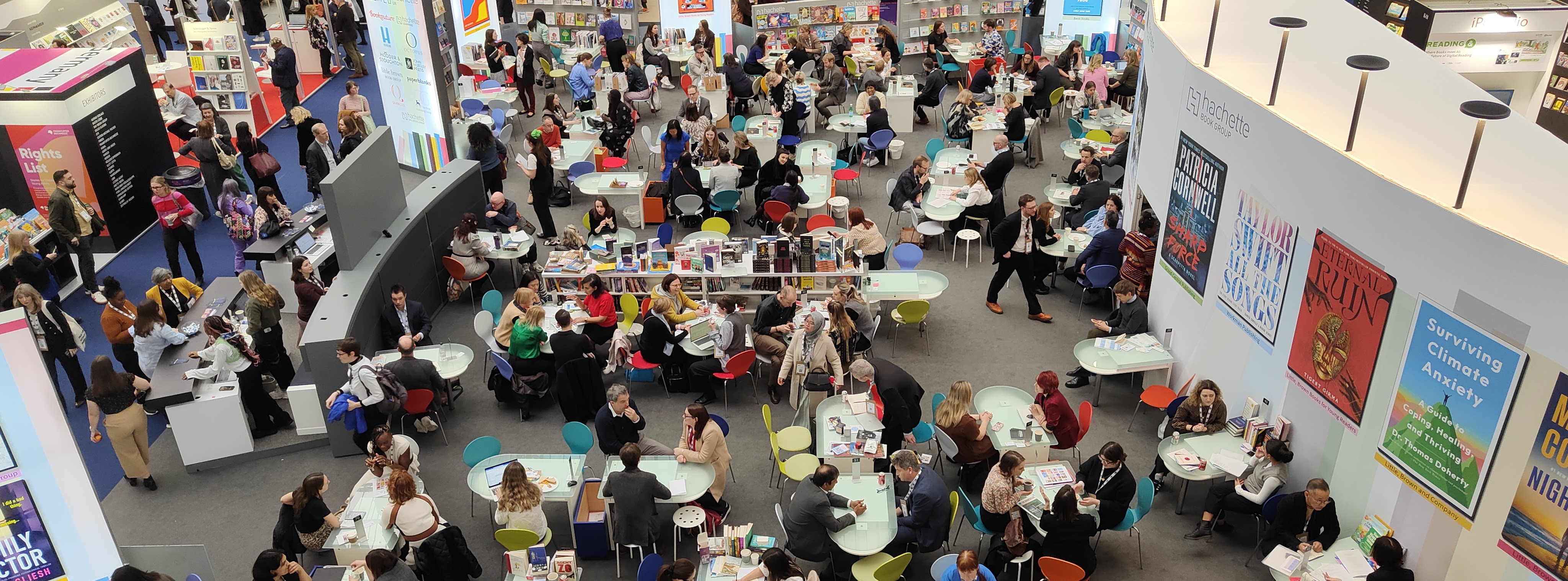 Aerial view of attendees engaging in discussions at tables in a busy area of the London Book Fair, with various books on display in the background.